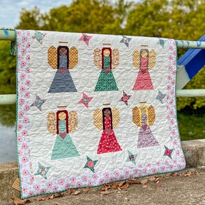 May include: A white quilt with a floral border and six angel figures in the center. The angels are made of patchwork fabric in various colors and patterns, including red, green, blue, and yellow. The quilt is hanging on a railing.