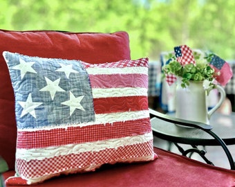 May include: A red and white patriotic cushion with a frayed edge and a blue and white American flag design. The cushion is sitting on a red chair.