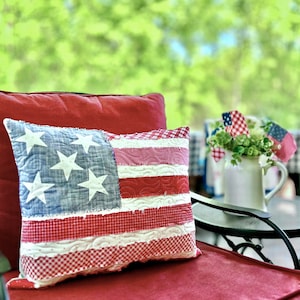 May include: A red and white patriotic pillow with a frayed edge and a blue and white American flag design. The pillow is sitting on a red chair.