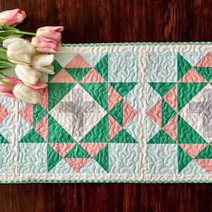 Puede incluir: Un camino de mesa acolchado con un patrón de cruz en tonos de verde, rosa y blanco. El camino de mesa tiene un borde de cuadros y está decorado con un ramo de tulipanes rosas y blancos.
