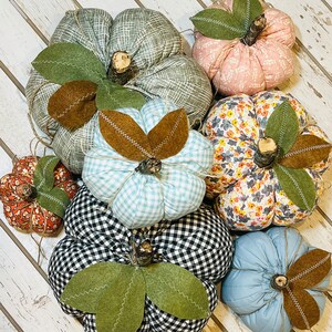 May include: A collection of six handmade fabric pumpkins in various patterns and colors, including blue gingham, black and white gingham, floral, pink, and green. The pumpkins are arranged on a white wooden surface.