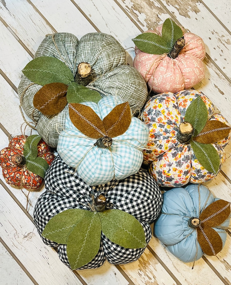 May include: A collection of six handmade fabric pumpkins in various patterns and colors, including blue gingham, black and white gingham, floral, pink, and green. The pumpkins are arranged on a white wooden surface.