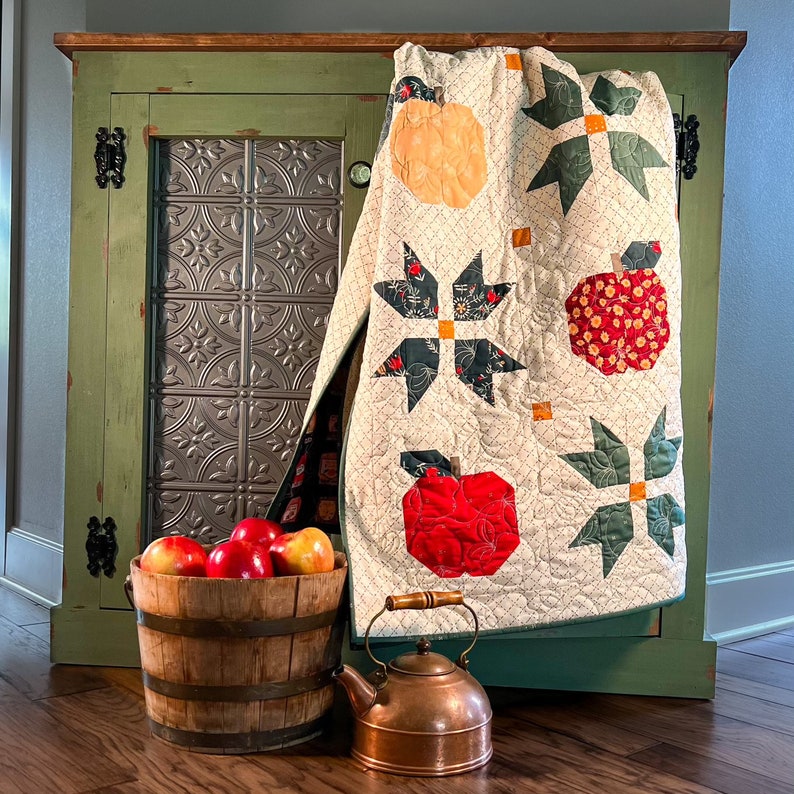 Puede incluir: Una colcha blanca con un dise&ntilde;o de manzanas verdes y rojas cuelga de un armario verde con una puerta de panel met&aacute;lico. Un cubo de madera lleno de manzanas rojas se encuentra delante del armario. Una tetera de cobre con asa se encuentra en el suelo.