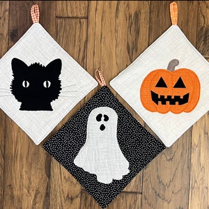 Pode incluir: Três pegadores de panela com tema de Halloween. Um apresenta um gato preto, outro um fantasma branco e outro uma abóbora laranja com um rosto de abóbora preto.