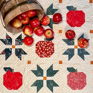 May include: A white quilt with a red, green, and white patchwork design. The quilt features a repeating pattern of apples and stars. The apples are red with a white outline and the stars are green with a white outline. There is a wooden bucket overflowing with red apples in the upper left corner of the image.