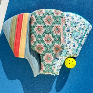 Peut inclure: Trois sacs en tissu matelassés avec des motifs floraux dans des tons de bleu, vert et rose. Les sacs sont disposés en forme de triangle avec une balle jaune au centre.