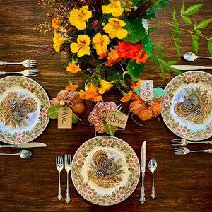 May include: A Thanksgiving table setting with three place settings, each with a white plate featuring a turkey design, silverware, and a place card. The table is decorated with a centerpiece of fall flowers and gourds.