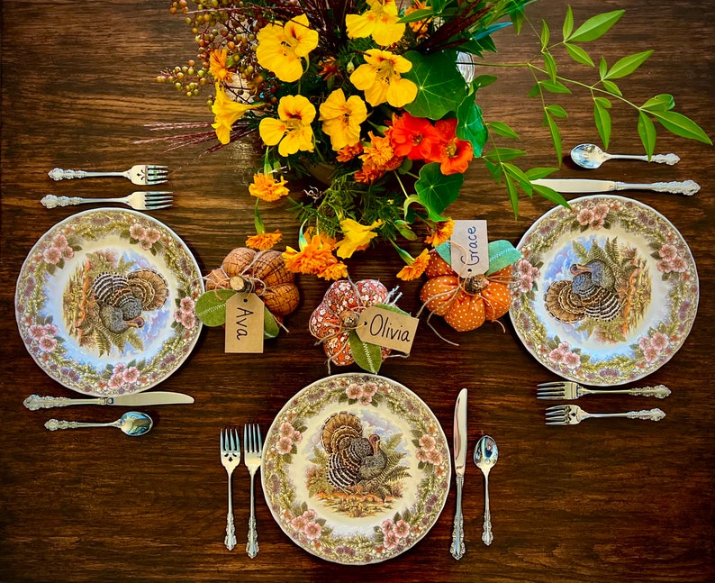 May include: A Thanksgiving table setting with three place settings, each with a white plate featuring a turkey design, silverware, and a place card. The table is decorated with a centerpiece of fall flowers and gourds.