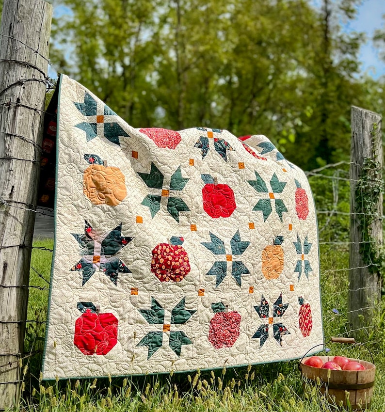 Puede incluir: Una colcha de color crema con un dise&ntilde;o de patchwork que presenta manzanas verdes, rojas y naranjas. La colcha est&aacute; colgada en una valla de madera con hierba verde y un fondo borroso de &aacute;rboles.
