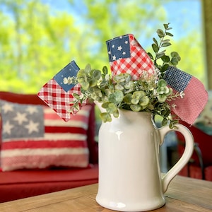 May include: A white ceramic pitcher filled with green foliage and red, white, and blue fabric flags. The pitcher is sitting on a wooden table.