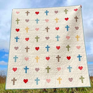 Peut inclure: Une courtepointe blanche avec un motif répétitif de croix et de cœurs. Les croix sont de différentes couleurs, notamment bleu, vert, marron et jaune. Les cœurs sont dans des tons de rose et de rouge.