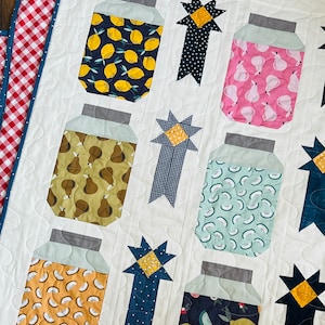 May include: A white quilt with a patchwork design featuring various jars with different fruit patterns. The jars are surrounded by blue and yellow stars with ribbons.