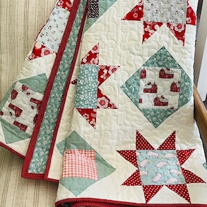 May include: A white quilt with a red and green patchwork design. The quilt features a variety of patterns, including stars, squares, and diamonds. The quilt is edged with a red trim.