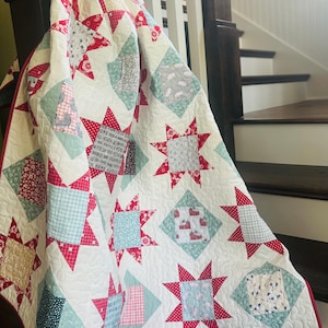 May include: A white quilt with a red and green star pattern. The quilt is made of various fabrics with different patterns, including plaid, polka dots, and floral prints. The quilt has the text "ONE GOOD THING A DAY" in the center.