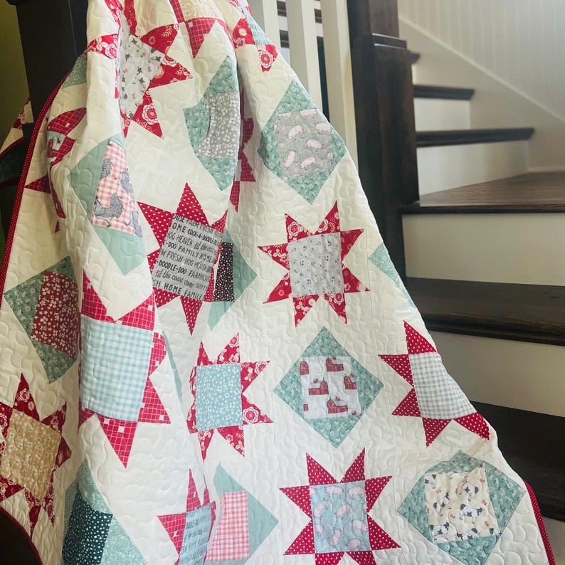 May include: A white quilt with a red and green star pattern. The quilt is made of various fabrics with different patterns, including plaid, polka dots, and floral prints. The quilt has the text "ONE GOOD THING A DAY" in the center.