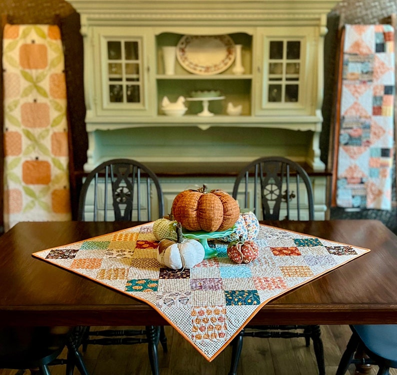 May include: A brown wooden table with a patchwork quilt table runner featuring a variety of autumn colours and patterns.  The table runner is decorated with four small fabric pumpkins, one of which is a larger orange pumpkin.