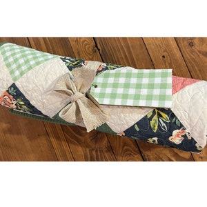 May include: A green, white and black quilted blanket with a floral pattern is rolled up and tied with a burlap bow. A green and white gingham gift tag is attached to the bow.
