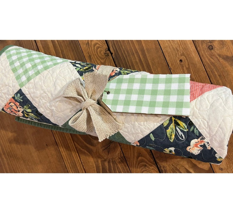 May include: A green, white and black quilted blanket with a floral pattern is rolled up and tied with a burlap bow. A green and white gingham gift tag is attached to the bow.