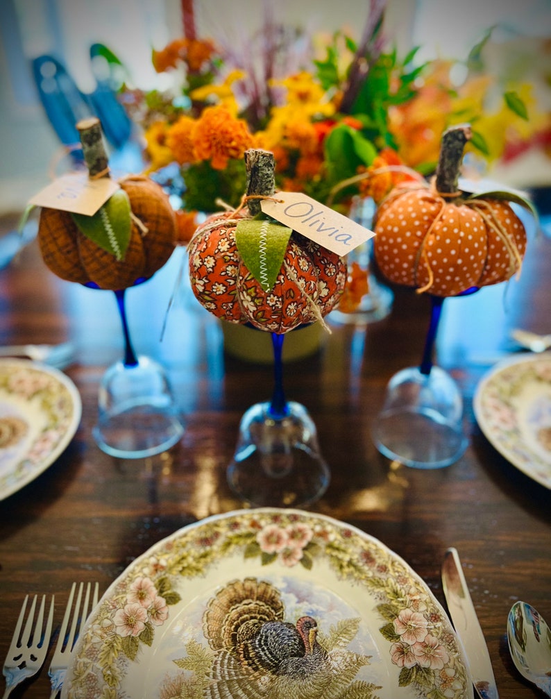 May include: A Thanksgiving table setting with three orange and brown fabric pumpkins with blue glass stems. The pumpkins have name tags with the names "Olivia" and another name that is not visible. The table is set with a white plate with a turkey design and a floral border.