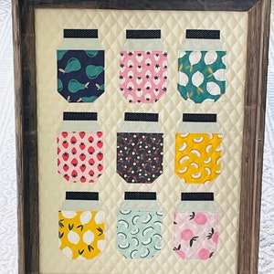 May include: A framed quilted wall art piece featuring nine mason jars with different fruit and floral patterns. The jars are arranged in three rows of three. The frame is brown wood.