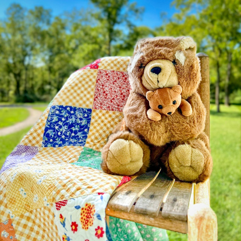 May include: A brown teddy bear holding a smaller brown teddy bear, sitting on a yellow and red patchwork quilt. The quilt is on a wooden chair.