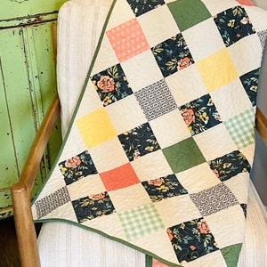 May include: A patchwork quilt featuring a mix of floral and geometric patterns in shades of green, yellow, pink, and black. The quilt is folded and draped over a chair with a light brown fabric.