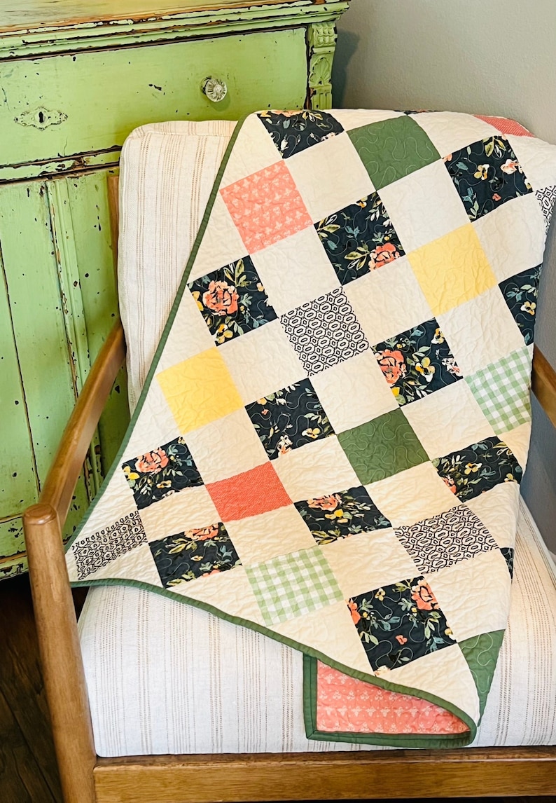 May include: A patchwork quilt featuring a mix of floral and geometric patterns in shades of green, yellow, pink, and black. The quilt is folded and draped over a chair with a light brown fabric.