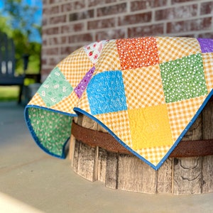 May include: A patchwork quilt featuring a mix of floral and gingham patterns in shades of yellow, orange, green, blue, and purple. The quilt is draped over a wooden barrel.