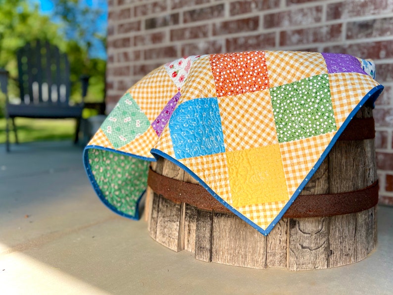 May include: A patchwork quilt featuring a mix of floral and gingham patterns in shades of yellow, orange, green, blue, and purple. The quilt is draped over a wooden barrel.