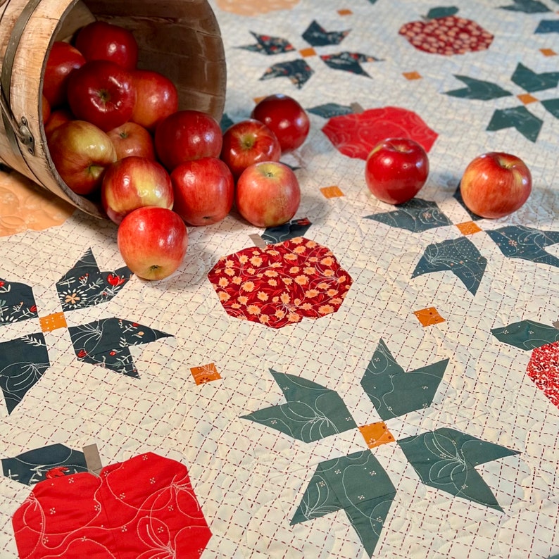 Puede incluir: Una colcha blanca con un dise&ntilde;o de manzanas rojas y verdes. La colcha est&aacute; hecha con una variedad de telas y tiene un patr&oacute;n acolchado. Un cubo de madera lleno de manzanas rojas se derrama sobre la colcha.