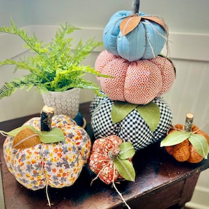 May include: A stack of five fabric pumpkins in various patterns and colors. The pumpkins are arranged from largest to smallest, with the largest pumpkin on the bottom and the smallest pumpkin on top. The pumpkins are decorated with a variety of fabrics, including floral, plaid, and polka dot patterns. The pumpkins are a great way to add a touch of fall decor to your home.