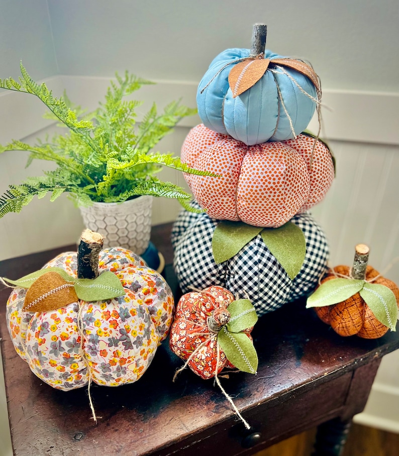 May include: A stack of five fabric pumpkins in various patterns and colors. The pumpkins are arranged from largest to smallest, with the largest pumpkin on the bottom and the smallest pumpkin on top. The pumpkins are decorated with a variety of fabrics, including floral, plaid, and polka dot patterns. The pumpkins are a great way to add a touch of fall decor to your home.