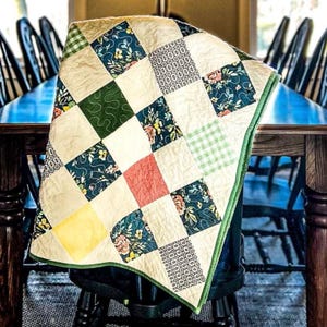 May include: A patchwork quilt featuring a mix of floral and geometric patterns in shades of green, blue, yellow, and red. The quilt is folded and draped over a chair.