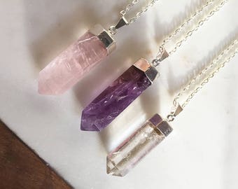 Puede incluir: Tres collares de cadena plateada con colgantes de cristal de punta. Los colgantes son de cuarzo rosa claro, morado y transparente.