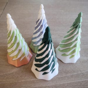 May include: Four decorative ceramic Christmas trees in various colours. The trees are white, green, blue, and orange, with white and green accents. The trees are arranged on a light brown wooden surface.