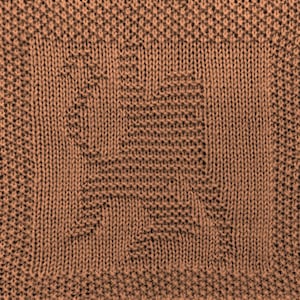 May include: A close-up of a brown knitted fabric with a textured pattern. The fabric is woven in a square shape with a repeating pattern.
