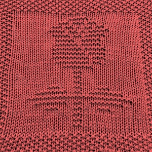 May include: Coral-coloured knitted square featuring a detailed tree design at the centre. The tree is created with a textured stitch, and the square has a decorative border. The overall texture is soft, suggesting a handmade item, possibly a blanket square.