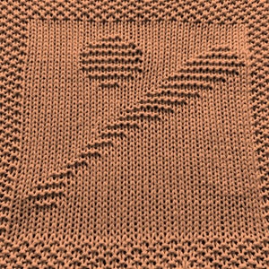 May include: A close-up of a knitted dishcloth in a light brown color. The cloth has a textured pattern with a diamond shape in the center.