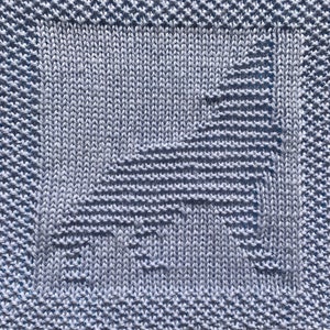 May include: Close-up of a knitted textile in shades of blue and white. The design features a central square with a geometric pattern, surrounded by a textured border. The knit stitches create a detailed and tactile surface, showcasing the craftsmanship of the piece.