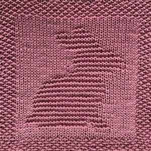 May include: A close-up of a knitted dishcloth in a deep pink colour. The cloth has a textured pattern with a geometric design in the centre.