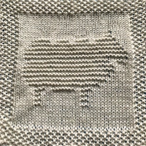 May include: A knitted square blanket in a light beige color. The blanket features a central design of a stylized animal, possibly a pig, within a rectangular border. The edges have a decorative knitted pattern.