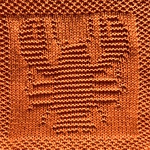 Puede incluir: Primer plano de un cuadrado tejido en un tono naranja cálido. El diseño presenta una forma de corazón central, creada con puntadas texturizadas, dentro de un cuadrado texturizado. El patrón de tejido es visible en toda la imagen.