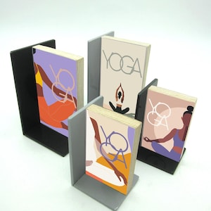 Peut inclure: Ensemble de serre-livres sur le thème du yoga. Chaque serre-livres maintient un petit livre avec le mot "YOGA" et une illustration de posture de yoga. Les serre-livres sont noirs et gris, avec des livres aux tons orange, violet et beige.