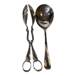 Op de afbeelding: Een set zilveren serveerbestek, bestaande uit een tang en een grote lepel. De tang heeft sierlijke handvatten en ringgrepen. De lepel heeft een afgeronde kom en een lange, slanke steel. De gebruiksvoorwerpen zijn reflecterend en lijken van metaal te zijn.