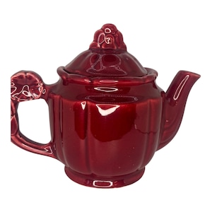May include: A small, glossy, burgundy-colored ceramic teapot. The teapot has a lid with a decorative floral design on top, a curved spout, and a handle with a floral accent. The teapot is designed with vertical ridges.