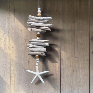 May include: A driftwood mobile, suspended by twine, featuring stacked driftwood pieces, wooden beads, and a white starfish. The backdrop is a wooden wall, with natural light casting shadows.