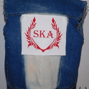 May include: A blue denim vest with a white patch sewn on the back. The patch features a red stylized wreath with the word "SKA" in red letters.