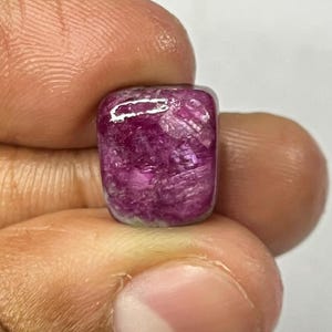 May include: A small, square-cut, polished gemstone with a deep magenta color. The gem is held between fingers against a white background, showcasing its vibrant hue and smooth surface. The stone's shape is a rounded square.
