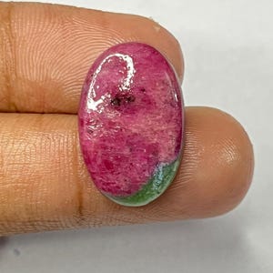 May include: Oval-shaped gemstone with a vibrant pink and red color, featuring a mix of textures and hues. The stone has a polished surface and a small area of green at one end. The gemstone is held between fingers.