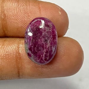 May include: An oval-shaped, polished gemstone with a deep magenta hue, featuring a marbled pattern of lighter pink and white veins. The gem is held between two fingers against a white background, showcasing its natural beauty.